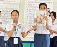A group of school students in white short-sleeved shirts and dark skirts or trousers stand in front of a white board giving a presentation. A young woman in the front left holds a microphone and speaks, gesturing with her left hand toward a classmate beside her. The young man holds up a printed photograph showing two decorated bamboo bottles nestled in green leaves. In the background, posters with text in English and Khmer script are visible, with one heading reading "Topic: Bamboo bottles for our community
