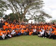 A wide shot of a large group of people posing together on a grassy lawn in front of a sprawling, leafy tree under an overcast sky. The majority of the group is wearing matching bright orange t-shirts with a graphic logo on the front, while a few individuals are in business or white shirts. Many people in the crowd are smiling and waving their hands at the camera. The individuals are arranged in rows, with those in the front kneeling or sitting on the grass, and those in the back standing.