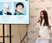 A young woman with light skin and long brown hair holding a microphone addresses an audience next to a screen displaying slide details and speaker headshots for "Education House"