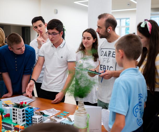 A teenage boy points to a colorful model of a city on a table while surrounded by several other teenagers and a man with a clipboard, all of whom are listening attentively