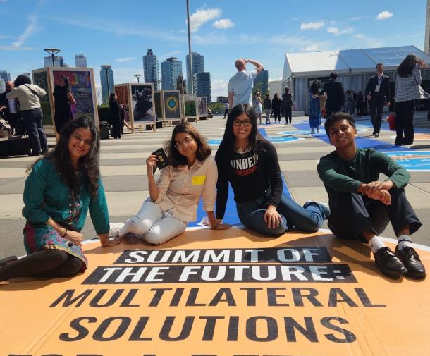 Four diverse young people sit on a yellow street mural that says Summit of the Future Multilateral Solutions with a blue sky and NYC skyline behind them