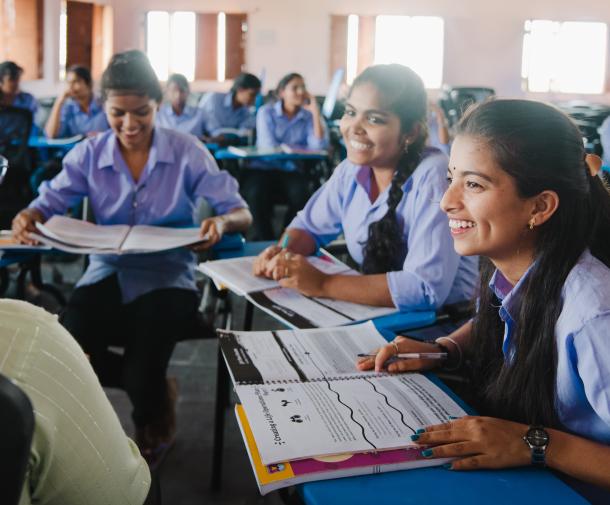 Three you girls sit in desks facing a teacher in a school, they are all smiling