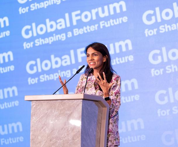 A young woman with long brown hair and tan skin stands at a podium in front of a background that says Global Forum for Shaping a Better Future
