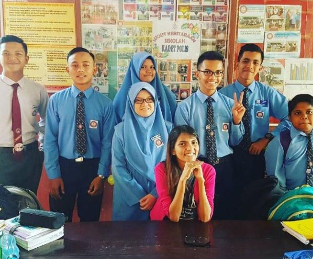 An Asian woman in a pink shirt smiles while seated at table surrounded by teenage students in blue uniforms