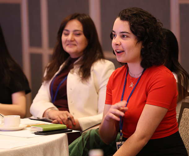 A young woman with shoulder-length brown curly hair and pale skin wearing a red shirt speaks while seated as others at the table watch and listen