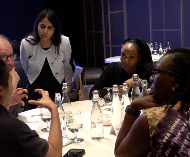 A woman with long brown hair wearing a white blazer leans over a table of diverse adults who appear to be in discussion