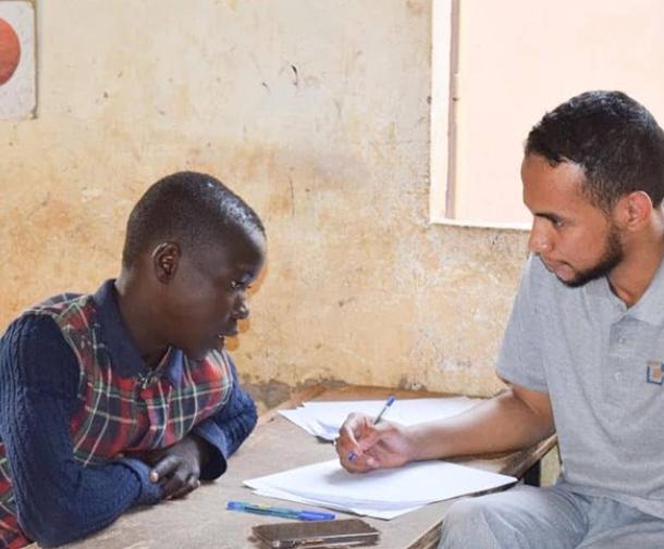 A black girl in a plaid school uniform and a young Black man in a grey Teach For Niger polo shirt look at a notebook together
