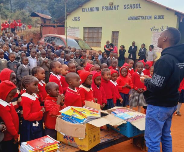A young Black man stands in front of a bench piled with workbooks and a large crowd of Black children in school uniforms outside of a school