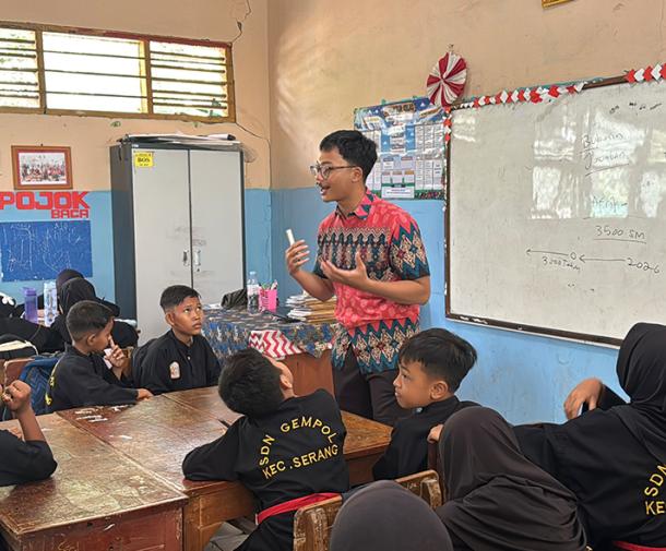 A teacher standing in front of a whiteboard, gesturing while presenting a lesson to students seated around tables in a classroom