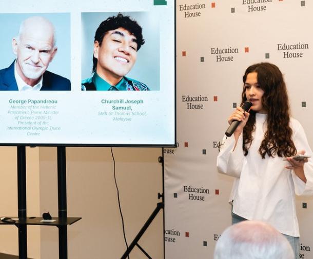 A young woman with light skin and long brown hair holding a microphone addresses an audience next to a screen displaying slide details and speaker headshots for "Education House"