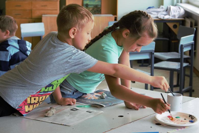 A boy in a grey shirt and a girl in a light green shirt dipping paintbrusheas in water and paint.