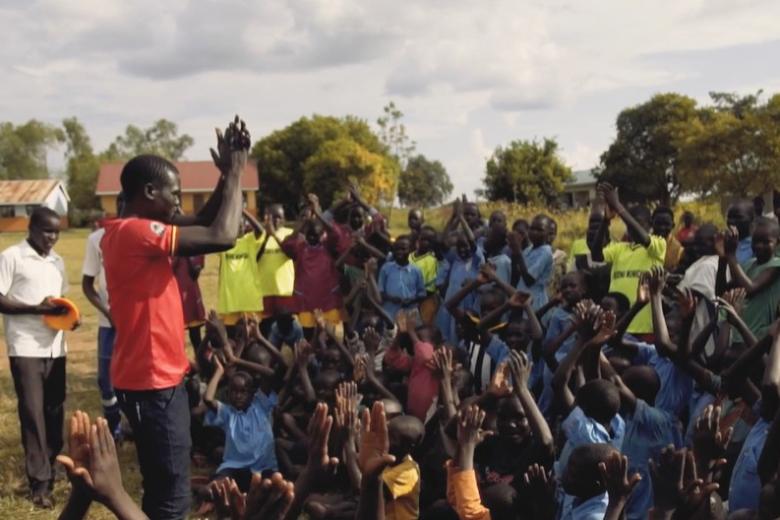 In a rural community in Africa a young man stands in front of a group of smiling children whose hands are raised