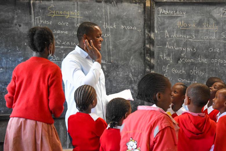 A Kenyan teacher stands at a chalkboard while young students in red uniforms cluster around him