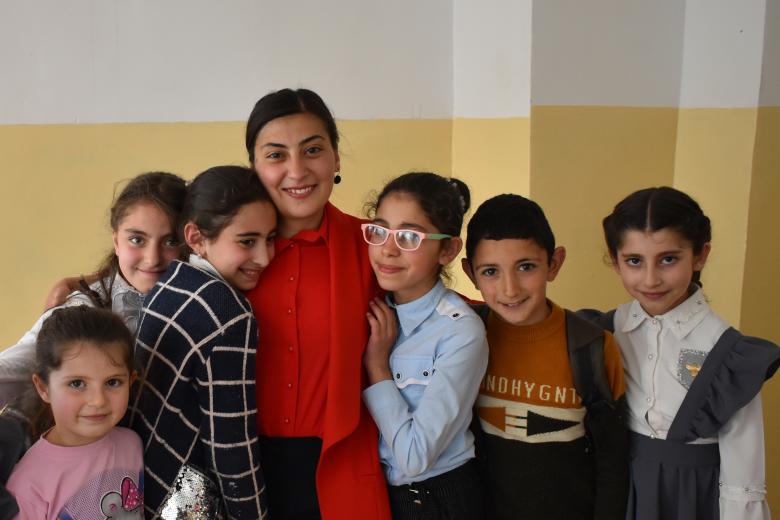 A female teacher in a red shirt and jacket has her arms around six students, one of whom rests her head on the teacher's shoulder