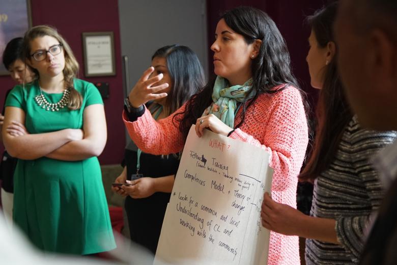 A White woman with brown hair speaks as she holds a large piece of paper. Five people of various ethnicities listen.