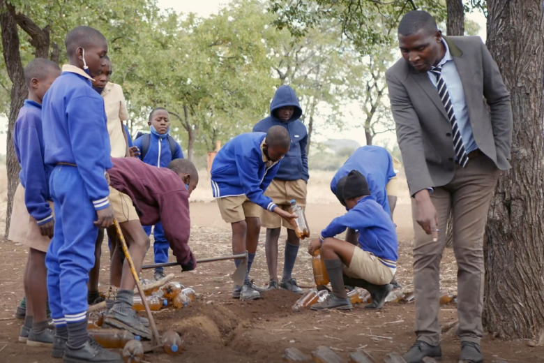 A young Black man in a jacket and tie points to the ground next to a tree as a group of students in blue school uniforms listens