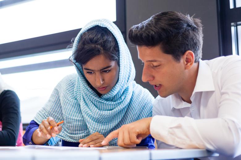 A young male teacher with tan skin and a teenage girl with tan skin, brown hair and a headscarf look at a worksheet together at a desk