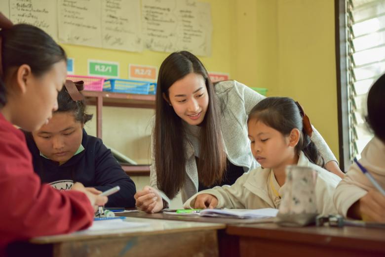 A young Asian woman with long hair leans over a table where three Asian girls are writing in workbooks