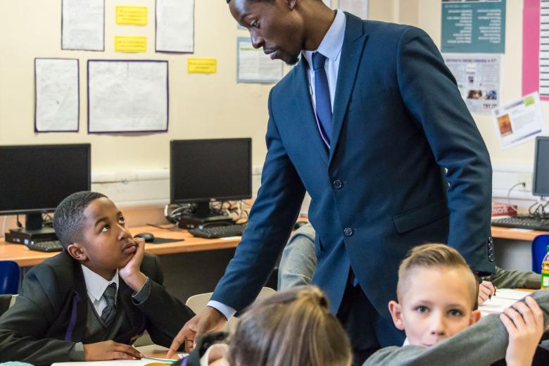 A young Black male teacher pints to a book on a desk as a black boy looks up at him and a white boy raises his hand