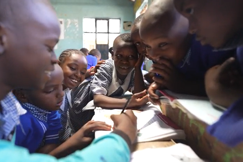 A group of young African boys leans over a shared desk smiling and writing in notebooks