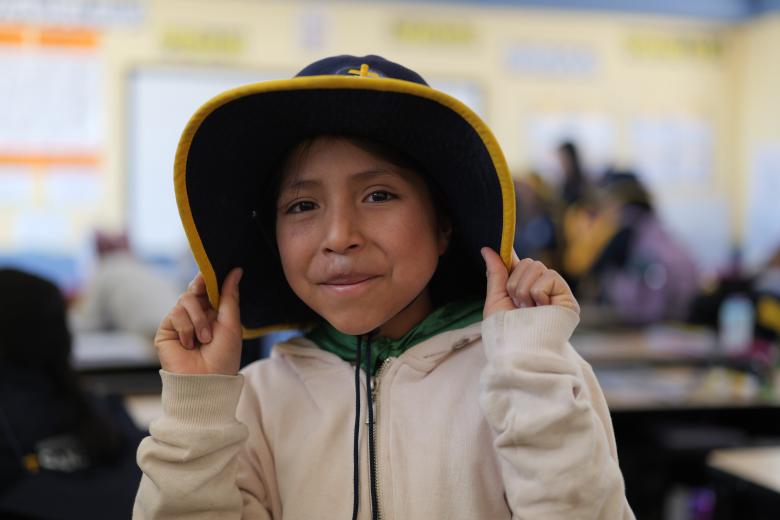 A girl with tan skin and brown hair in an off-white hooded sweatshirt pulls the sides of a black brimmed hat around her face and smiles