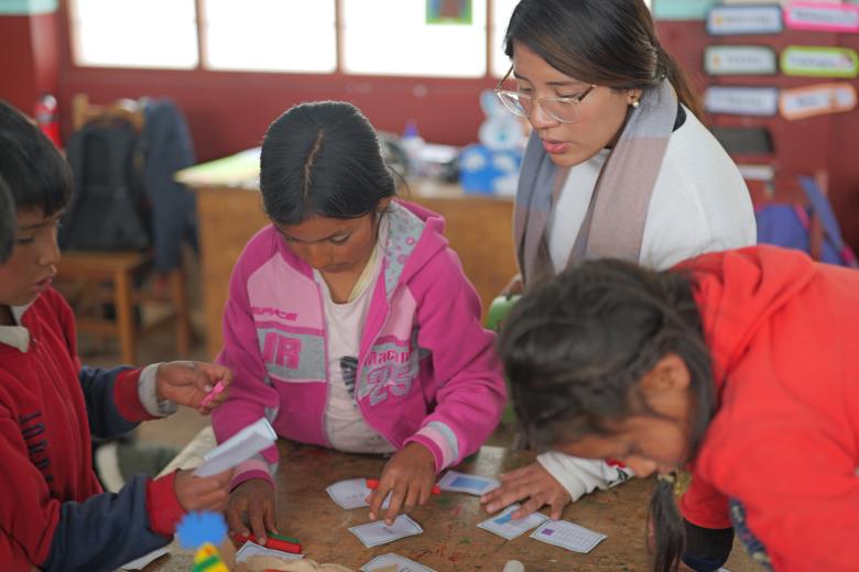 A female teacher with tan skin and brown hair pulled back leads over a table where two young girls and two young boys with tan skin and brown hair work on a project that involves cards