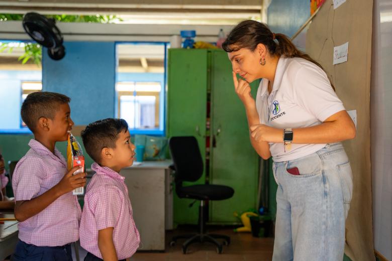 A female teacher with light tan skin & brown hair in a ponytail stands pointing to her nose and facing two young boys with tan skin and dark hair