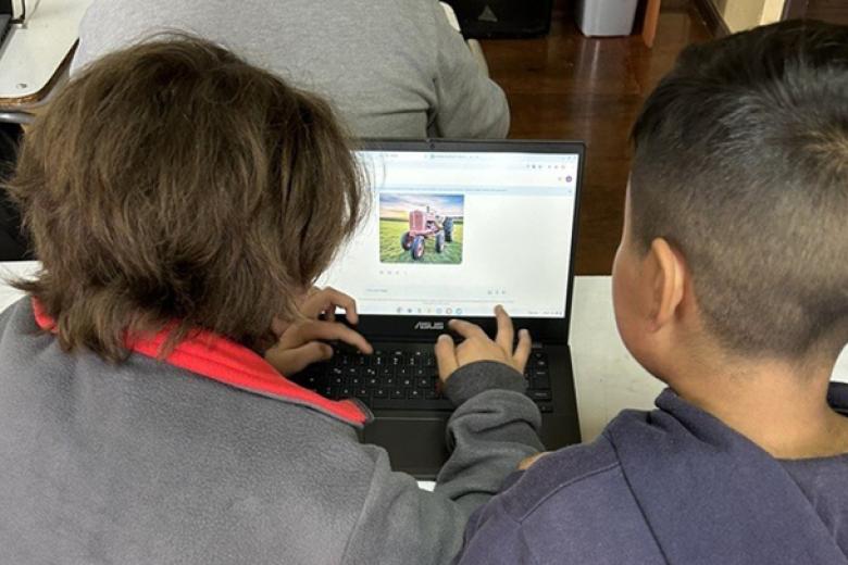 Seen from behind, the heads of two young children with brown hair and tan skin sharing a laptop computer