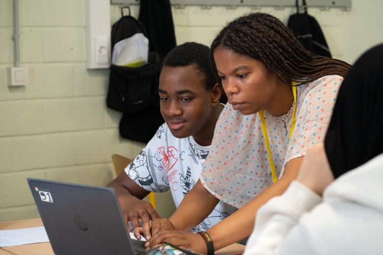 A Black female teacher types on a laptop computer next to a Black male teenager