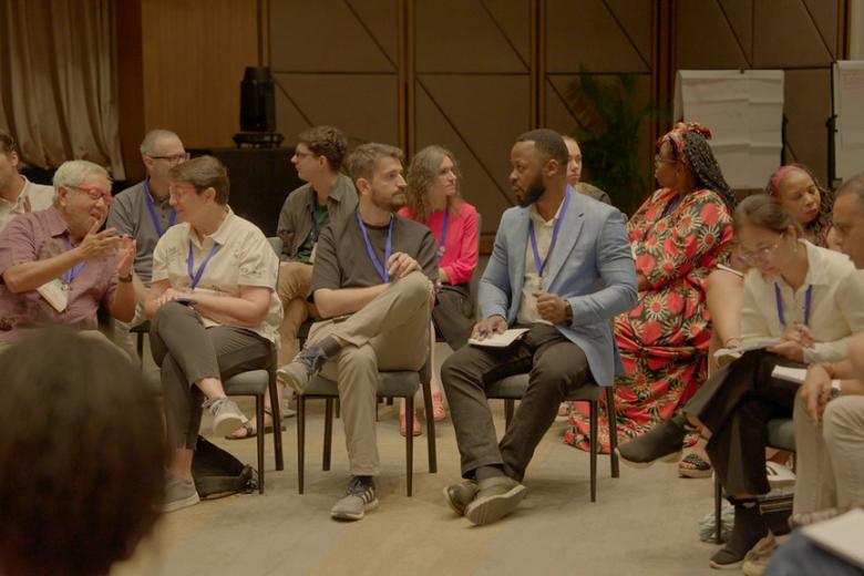A Black man in a light blue blazer talks to a white man in a blue shirt while seated among a diverse group of adults on chairs in what looks like a conference room