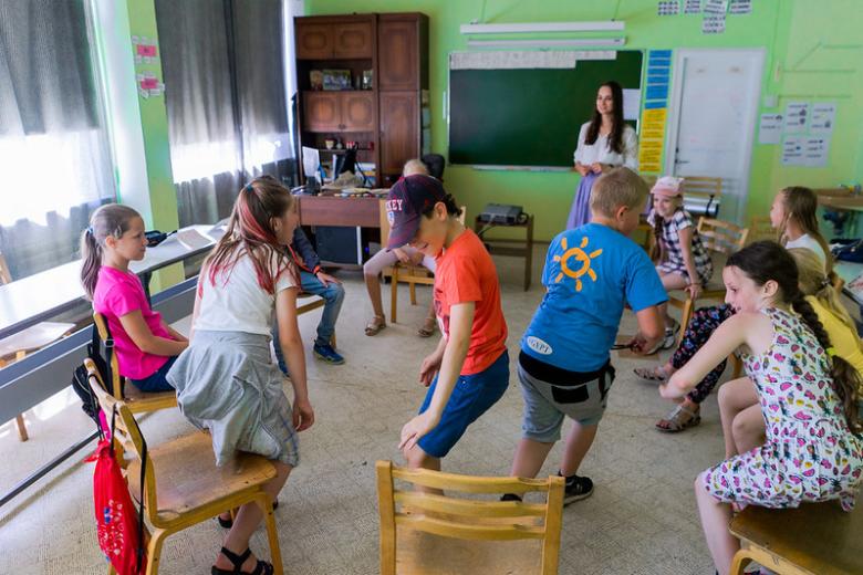 Children in a circle of chairs for a game in a classroom with a teacher.