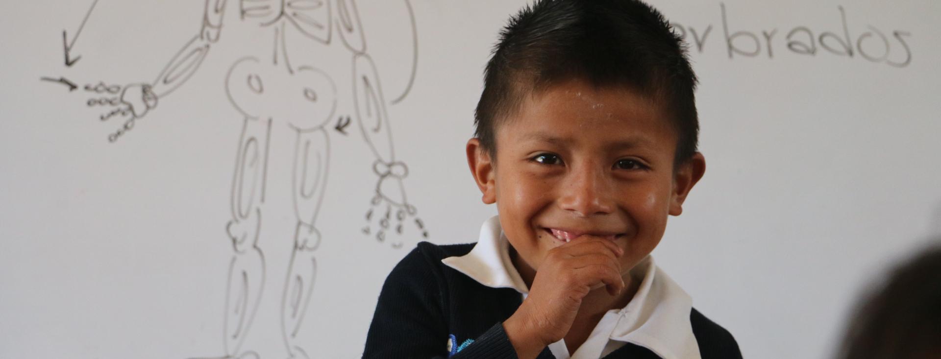 A close up of a young boy smiling broadly in front of a white board with a skeleton drawn on it and a word in Spanish that is partially obsured by the boy's head