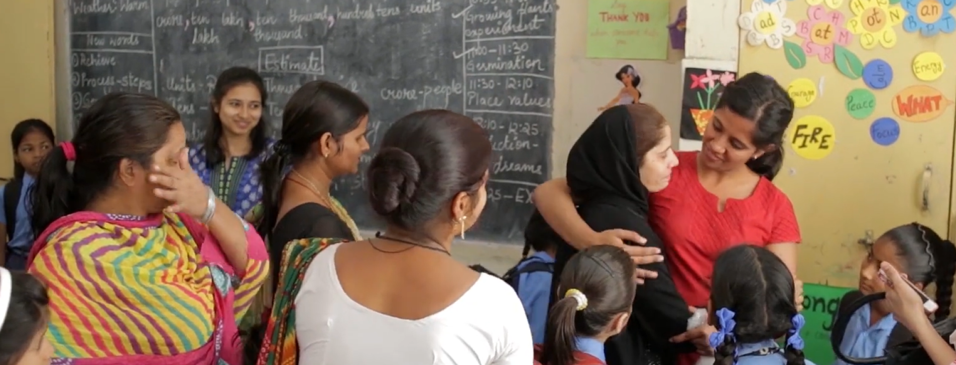 In a classroom, a young Indian woman in a red dress has her arm around another woman with a head scarf, surrounded by other women and girls in school uniform