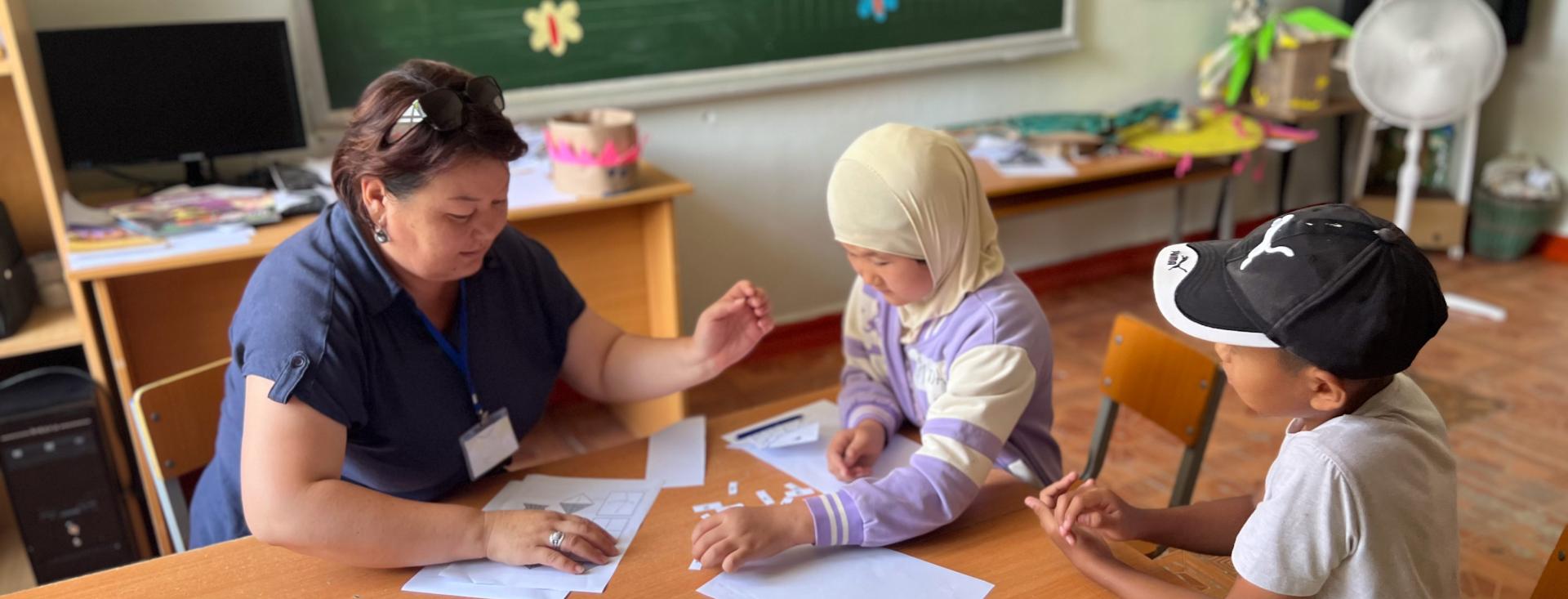 A young female teacher in a blue top shows worksheets to two young students at a table, one wearing a headscarf and the other a baseball cap