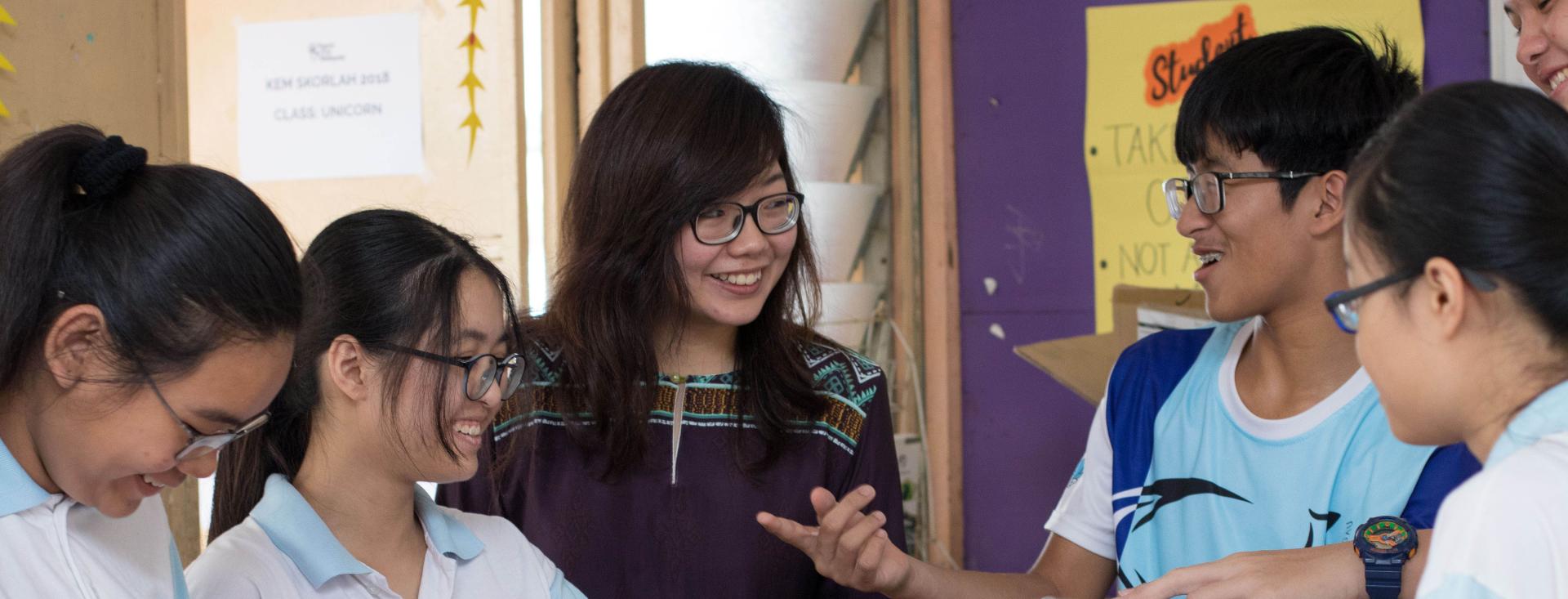 A young Asian female teacher is surrounded by 4 smiling teenage Asian students, 3 girls and a boys