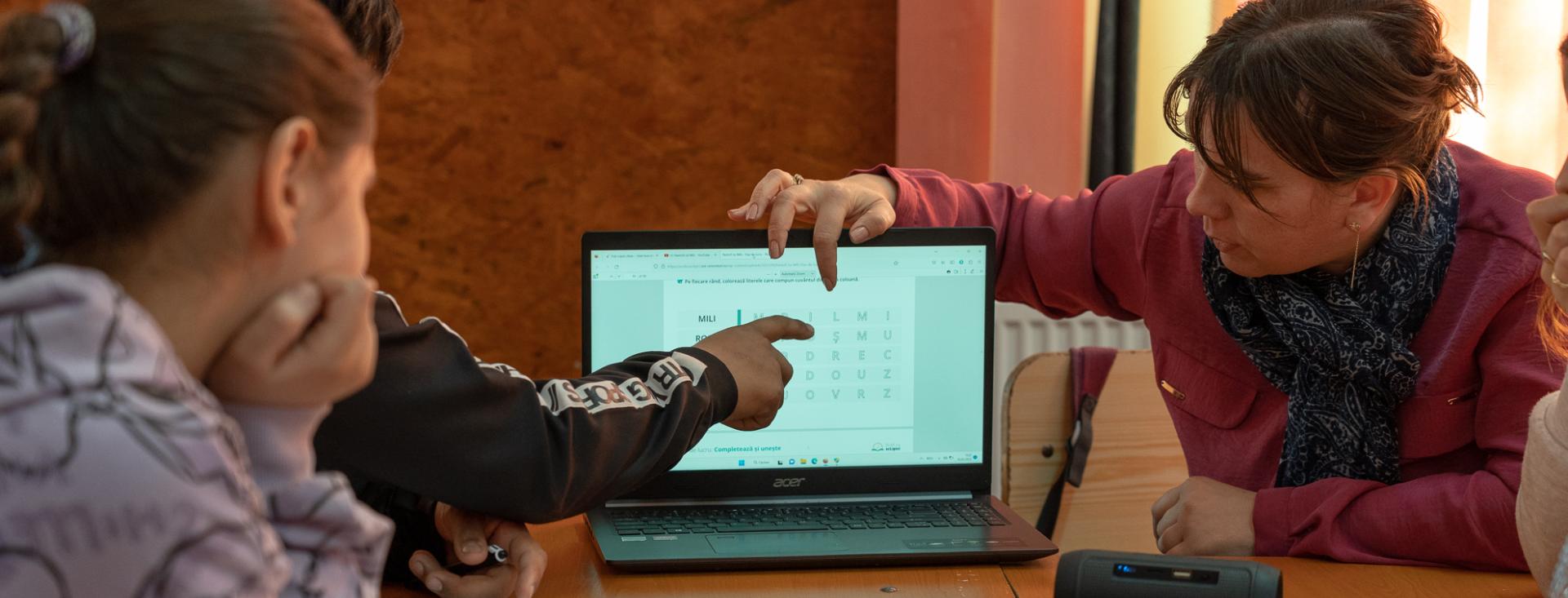A female teacher on one side of a laptop points to the screen while a male student on the other side also points to the screen and a female student watches