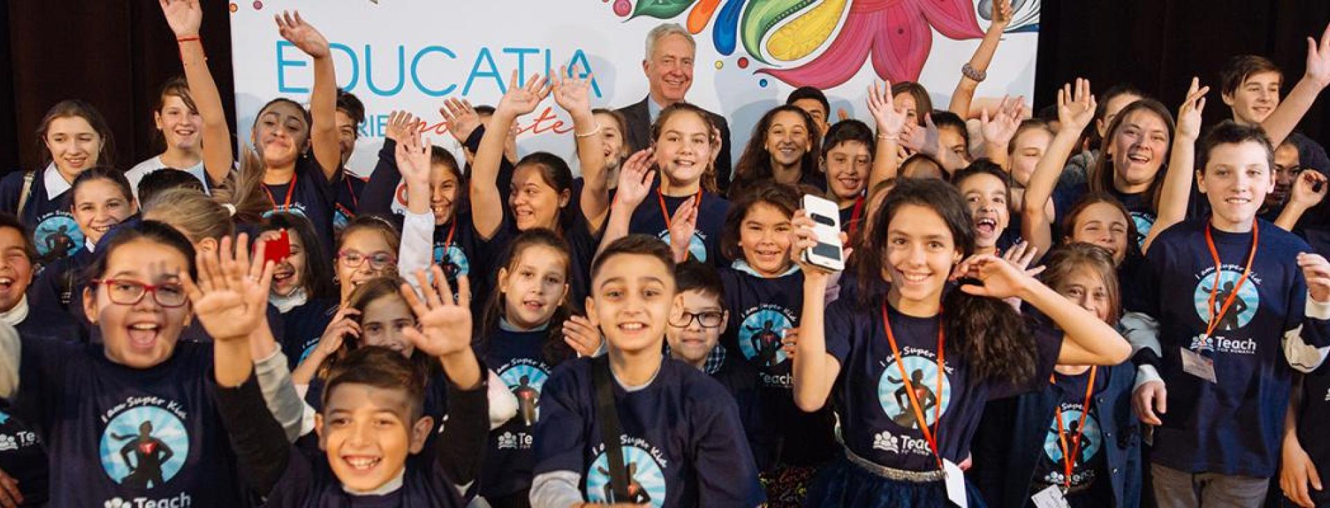A crowd of smiling children in matching "Teach for Romania" t-shirts cheer, wave, and raise their hands on stage at an event.