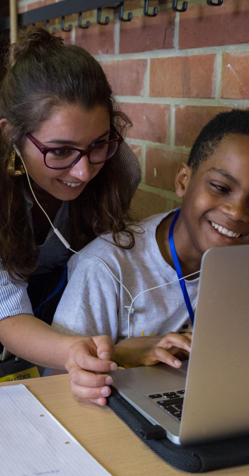 A young woman leans over two boys who are using a Macbook