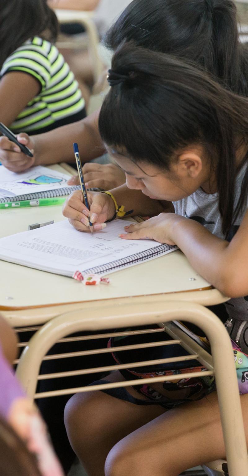 A young teen girl with a high ponytail writes in a notebook on her desk surrounded by classmates working at their desks