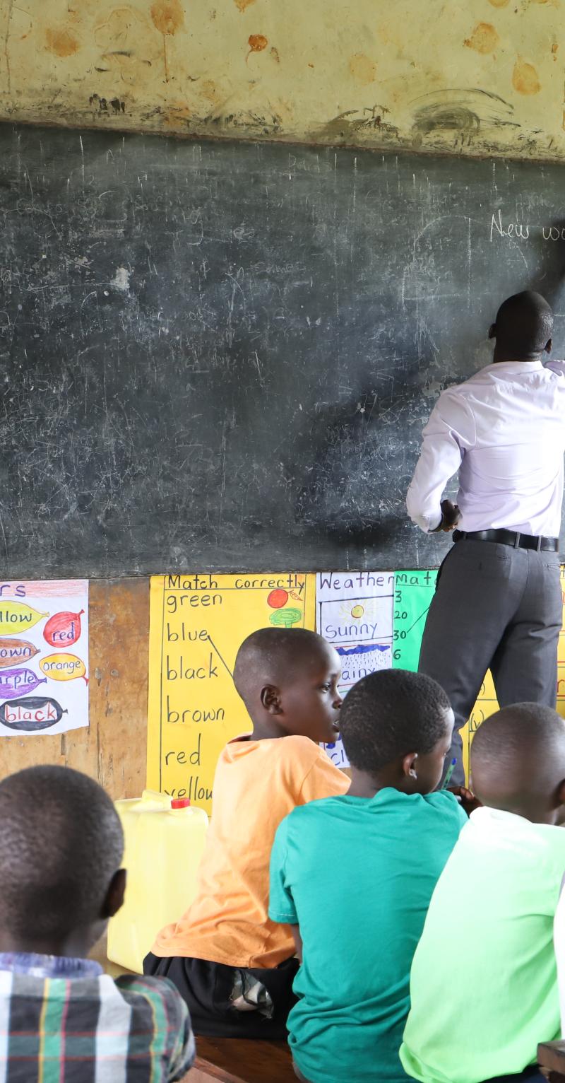 A young Black man in a white shirt writes on a chalk board with his back to a class of children seated in desks