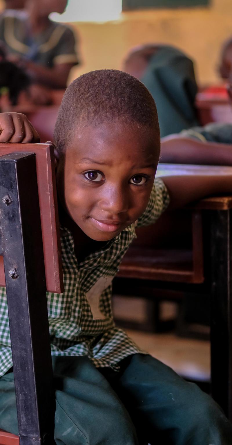 A smiling young black boy peers out from behind a wooden desk 