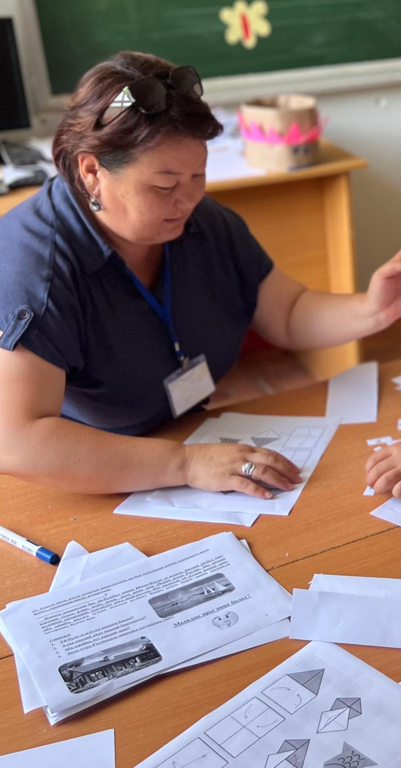 A young female teacher in a blue top shows worksheets to two young students at a table, one wearing a headscarf and the other a baseball cap