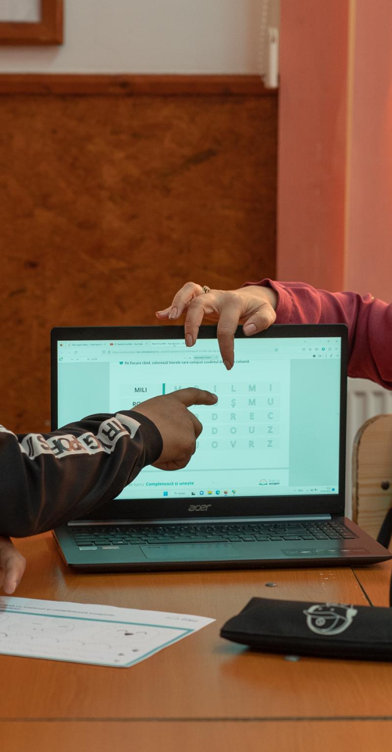 A female teacher on one side of a laptop points to the screen while a male student on the other side also points to the screen and a female student watches