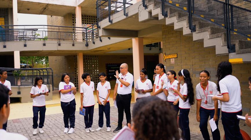 A young male teacher in a white shirt with a shaved head stands in a courtyard speaking, surrounded by teenage students also in white shirts