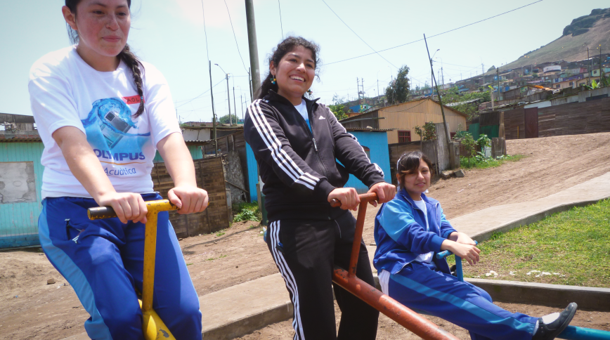 A young woman in a black track suit smiles as she sits on top of a see saw with smiling girls in blue track suits on either side