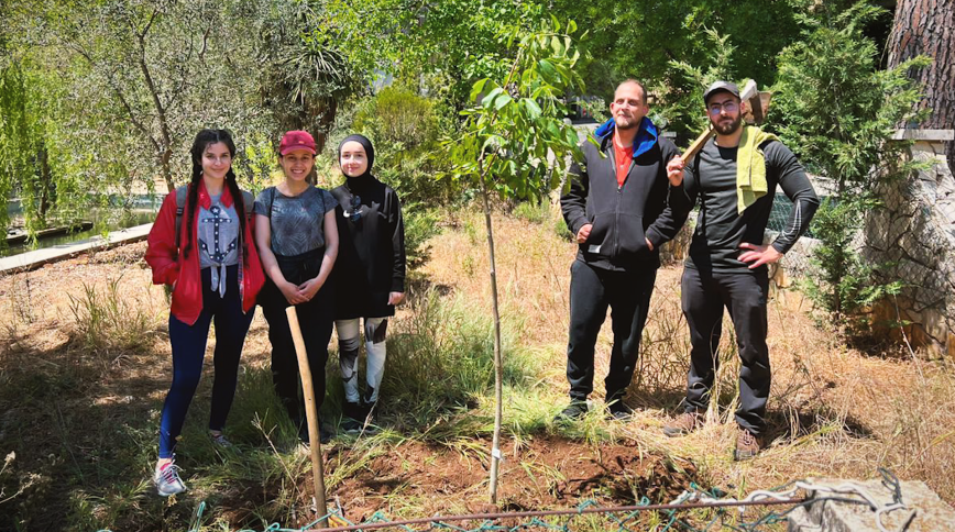 Two bearded men holding shovels and three young teen girls stand near two freshly planted saplings