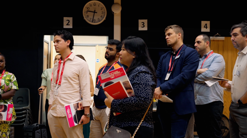 A diverse group of adults holding notebooks and wearing nametags stands in a school gym attentively looking out of frame as if someone is speaking to them