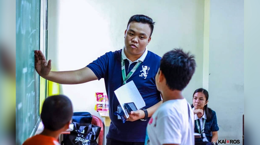 A young Asian man in a blue polo shirt speaks to a boy while pointing to a black board