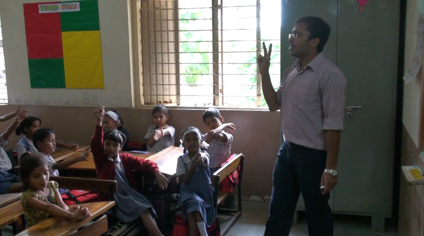 A young Indian male teacher stands in front of his class of young students holding up two fingers