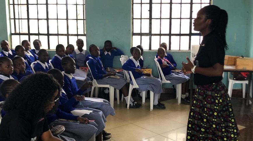 A young Black female teacher stands in front of a class of Kenyan students in school uniforms